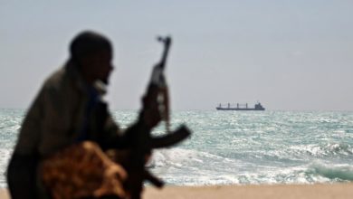 Man with gun looks across ocean at cargo ship.