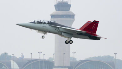 A T-7A Red Hawk is seen taking off from an airport. The words "US Air Force" are painted on its body, on the side of the cockpit. Its red tails have the number 7002 painted in black. An airport tower is seen in the background.