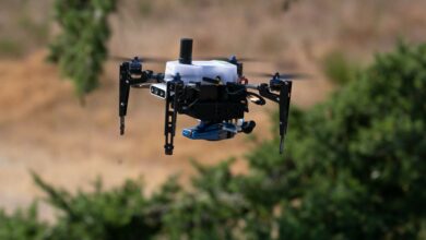 A small hovering drone is front and center in the picture. In the blurred out background, some greenery are seen in an otherwise rocky/sandy area.