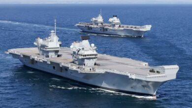HMS Prince of Wales and HMS Queen Elizabeth pictured at sea. The water is a deep blue, and the cloudy sky serves as the background.