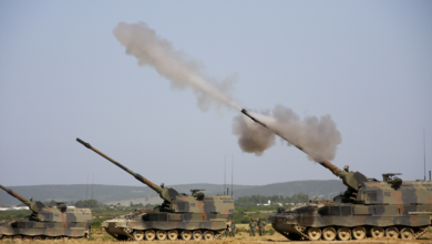 Three tanks, facing left, all have its main gun pointed upwards at a 45-degree angle. The rightmost tank, which is also closest, is in the process of firing ammunition. Smoke is seen shooting out of the gun, with the blurred ammunition in front.