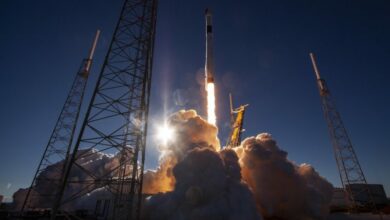 A rocket begins its launch. A huge cloud of smoke is seen below from it as it rises up, a red glow coming from its underside. The dark background is the night sky.