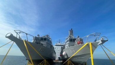 Fast-attack interdiction crafts (FAIC) PG-906 and PG-907 are seen docked in a port, both with two yellow ropes from the ground each tied over their hulls. Their names are painted in white on the side of their bodies. The blue sky acts as a background, with the port's blue-green water peeking on the lower back of the image.