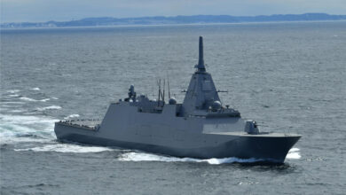 JS Mogami (FFM-1) is seen sailing in open water. A small strip of land serves as the distant background of the image. The ship's hull is colored grey, and the water is a dull, grayish blue.