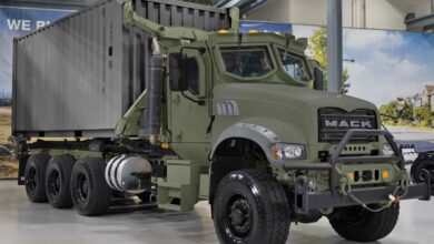 A Mack truck is seen on display at what appears to be a warehouse. It's colored dark green and has the 'MACK' logo in front of the truck.
