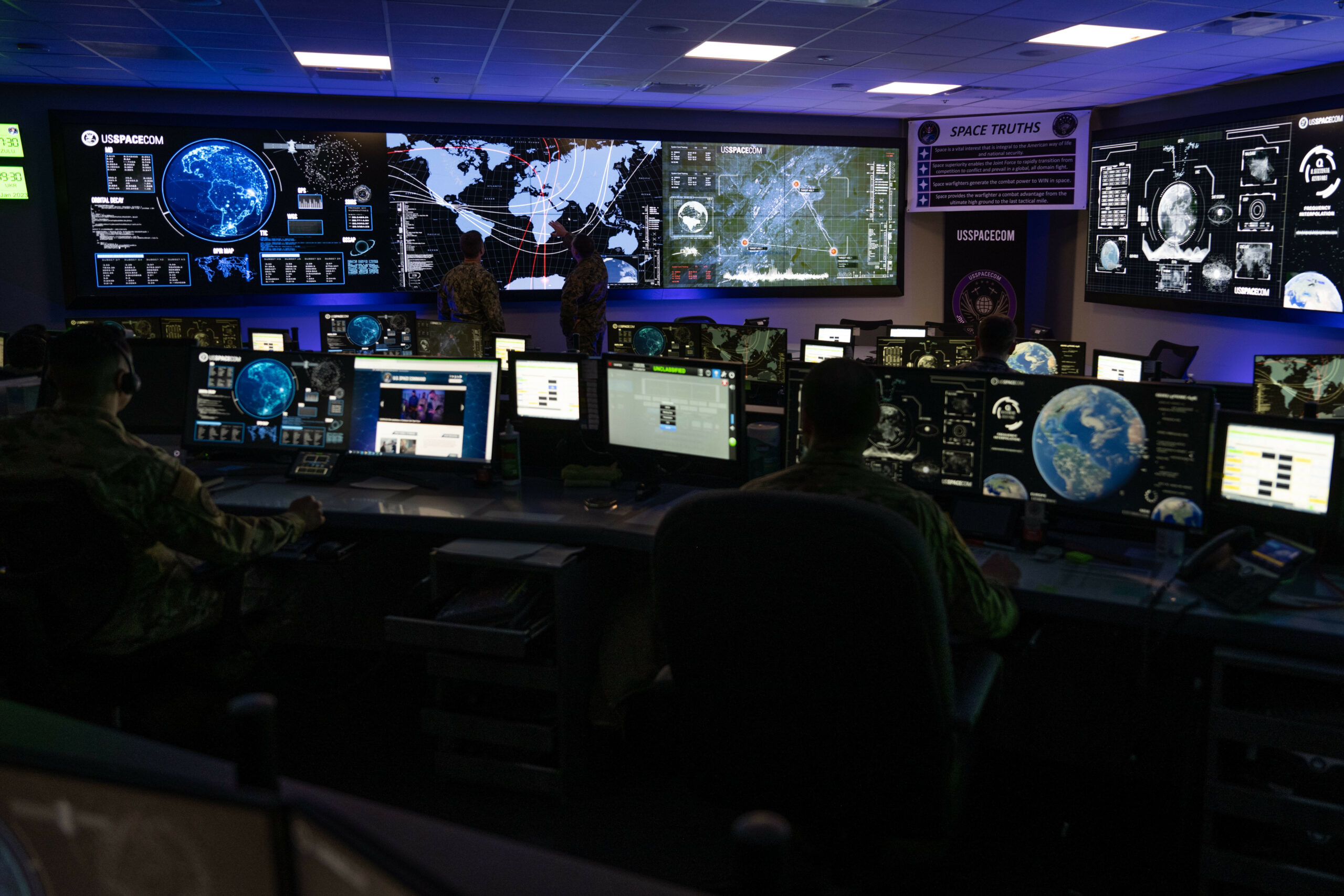 The dimly-lit room of the US Space Command Joint Operations Center is seen in the photo. The far left wall has three large monitors mounted on it, while the far right wall has one visible. The room is full of desks with uniformed personnel working on their computers. Most of the screens, including the walls' TVs', are displaying various interfaces that include maps and graphs.