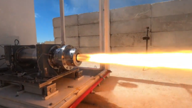 A rocket engine or launcher is set up in what appears to be an outdoor testing facility. A stream of bright yellow-orange jet flames is seen jutting out of its main exhaust. In the background, a concrete wall is seen, with the blue sky visible farther back.