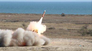 A missile from the Patriot missile system takes off in the middle of a field. The red missile is seen beginning to blast off, leaving a trail of smoke behind and a jet of white-orange flames. The background is a calm and blue body of water that stretches to the horizon, and a light-blue, cloudless sky.
