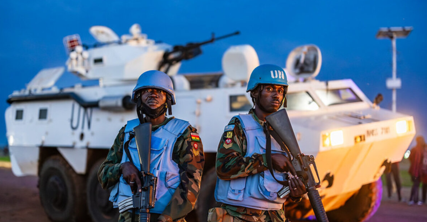 Two uniformed soldiers stand front and center of the image, staring straight. They both hold automatic rifles, pointed down. They are wearing a blue helmets and vests. The soldier on the right has 'UN' written in white in front of his helmet. Underneath their vests, they have a camouflage uniform on. A white tank with 'UN' written on its body in black is seen stationed in the background. Its orange headlights are on. The background is a dark blue sky.