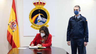 Span's Defense Minister Margarita Robles is seen signing a document inside what appears to be a conference room. An office personnel is seen standing by her left. The Spanish Air and Space Force emblem is displayed on the background, and the Spanish flag is positioned to her right.