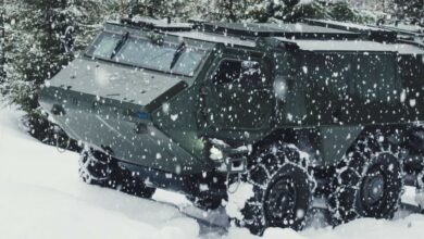 A dark green Patria 6x6 armored vehicle is seen traversing a snowy field while it's snowing. Trees can be seen on the background, covered in white snow. The vehicle's wheels are fitted with snow chains to help it have better grip and control on the ground.