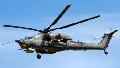 A Mil Mi-28 helicopter is seen hovering on the clear blue sky. On its gray-green hull, the number "03" is painted in yellow, as well as two red stars and the unit name "RF-93275" in white. Some characters on the Russian alphabet are also painted in black.