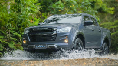 A gray Isuzu D-Max is seen being driven across a shallow pool of water in what appears to be an off-road location, with uneven ground and a background of green-leaved trees. The clear-white water around the truck's wheels are seen splashing towards all directions.