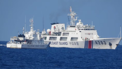 A white Philippine Coast Guard ship is stationed in the waters of the West Philippine Sea, facing a larger Chinese Coast Guard ship. The sea is a deep blue color, and the sky is a dull blue.