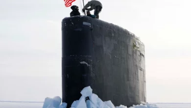 The USS Hampton's sail is seen breaking through the Arctic's massive ice flats. The submarine's sail is the only visible part of the vehicle. The ship is painted black. On top of the sail, three individuals are attaching a flag of the US on what appears to be a makeshift flag pole. The background is a completely white landscape and sky blending seamlessly together.