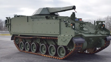 The Armored Multi-Purpose Vehicle Mortar Carrier prototype is seen in a large parking area. The tracked armored vehicle has a turret jutting out from its body. The entire vehicle is painted dark green.