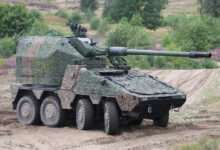 A KNDS Remote Controlled Howitzer 155 mm is seen on a grassy and sandy area. The eight-wheeled vehicle is painted with textured green and brown camouflage. Its turret is jutting forwards from the back. In the ground, trees and bushes grow scattered.