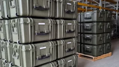 Dozens of military equipment boxes are seen stacked on top of each other in what appears to be a warehouse. They are supported underneath by wooden crates.