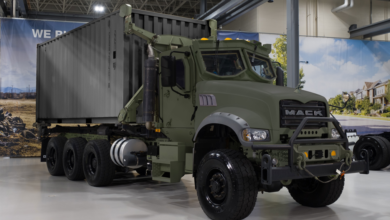 A Mack Common Tactical Truck is seen on display at what appears to be an arms/miitary exhibit. The 8-wheel truck is painted deep green. It has a cargo container in the back, painted a dull gray.