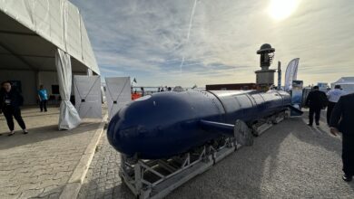 Elta Systems' Blue Whale autonomous submarine. The submarine is seen propped up on display at what appears to be an outdoor expo close to a body of water, slightly visible in the background. The submarine's slender body is painted a deep blue. At least nine individuals are seen scattered across the expo's grounds.