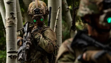 Two soldiers are seen wearing Enhanced Night Vision Goggle-Binoculars (ENVG-Bs). One soldier is on the far right foreground, out of focus. The other one is on the left side of the image. Both are wearing camouflage to match the wooded environment. The binoculars are strapped to their helmets, and its eyepiece are glowing bright green.