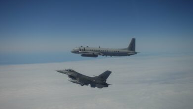 A Portuguese F-16 fighter jet escorting a Russian aircraft out of NATO's airspace above the clouds. Both planes are painted gray. The background is a sea of white clouds and the clear blue sky above.