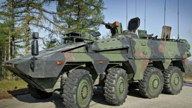 A Boxer armored fighting vehicle is seen parked in a cemented area in front of a green, tree-filled field. The eight-wheel armored vehicle is painted in green, brown, and black camouflage. A mounted gun by its hatch above is seen pointing forward. The background is a clear blue sky.