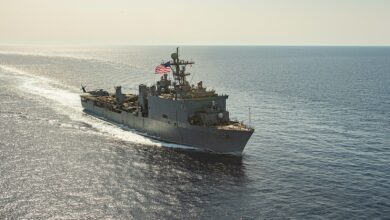 The dock landing ship USS Carter Hall (LSD-50) is seen sailing in calm waters. The sun, out of frame, shines a birght yellow-orange hue on the waters and the ship. The ship is painted gray, and has the US flag flying at full mast.