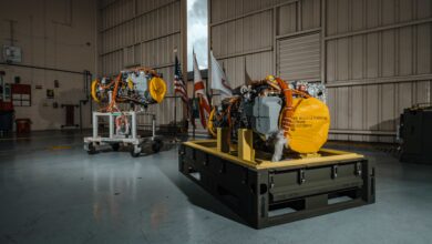 Two General Electric (GE) Aerospace T901 engines are displayed inside what appears to be a big warehouse or aircraft hangar. Wires, exposed metal, and other parts of machinery make up the engines.