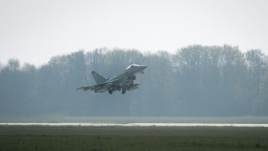 A UK Royal Air Force Typhoon jet takes off. The aircraft is colored gray, with 350 painted on its rudder. The background, partially covered in fog, appears to be a forested area.