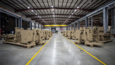 About eight units of Indirect Fires Protection Capability Increment 2 (IFPC Inc 2) ground-based systems are seen lined up in two rows in a large facility that appears to be a warehouse or a factory. The large units are colored light brown.