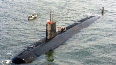 USS Toledo (SSN 769) is seen partially submerged in deep, blue-green waters. About six crewmen are seen atop the submarine. A small patrol boat is seen beside the submarine, on the upper left side of the image, with four people on board.