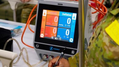 A medical equipment with a built-in screen that shows multiple values on the screen. Some of it appears to be measuring blood pressure. Its display is colored orange on the left and blue on the rigth, with both sides displaying the word "Deflated" most prominently. Four small tubes are connected to it, two orange tubes on the left and two clear ones below the screen. It appears to be resting on a table with a flat white surface. On the foreground, an arm of a uniformed personnel wearing green and brown camo fatigues can be seen writing something out of frame with a black pen.