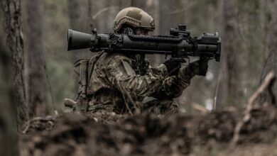 A soldier is seen in the center of the image carrying a Saab Carl-Gustaf Rifle on his shoulder. The rifle is painted black, and the soldier is wearing brown-green camouflage, blending in with the forest's colors.
