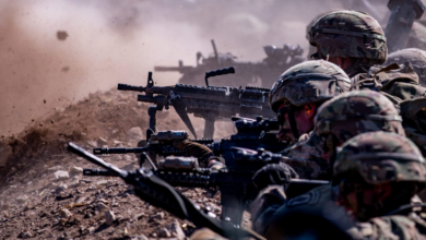 Four camo-wearing soldiers are seen positioned in a trench, pointing guns toward a target that is out of frame.