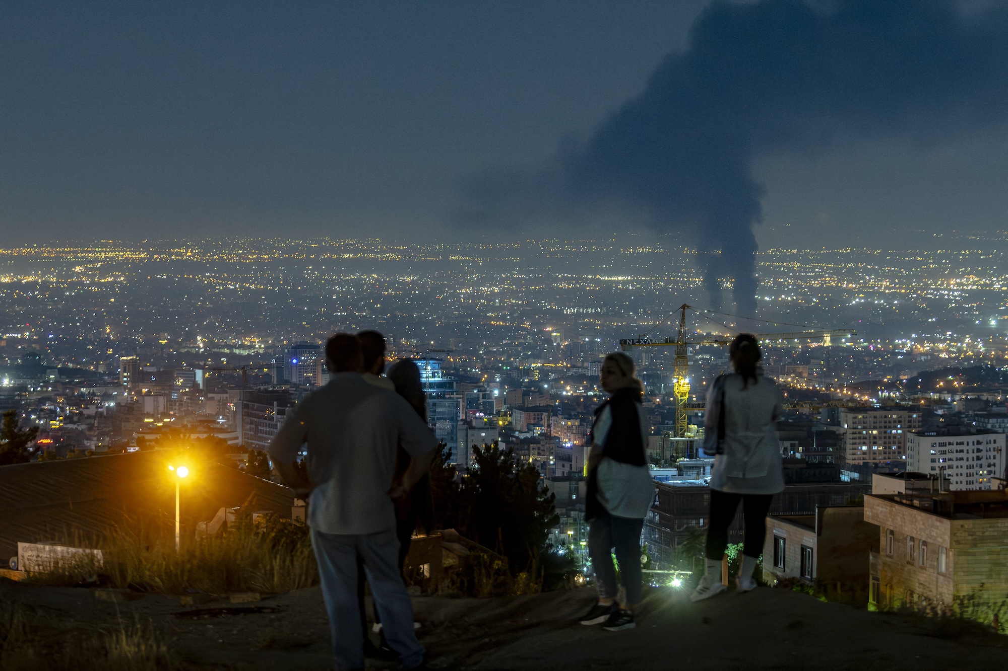 People gather on a hill to watch smoke rising in the distance from an Israeli airstrike in Tehran, Iran, on June 14, 2025
