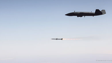 An MQ-28 Ghost Bat flies high above the ocean as it launches a missile, captured mid-flight with the weapon’s exhaust trail glowing against the clear sky