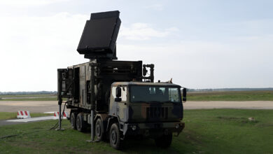 A camouflaged mobile military radar truck with its large flat-panel antenna raised and deployed on a grassy field near an airfield runway.