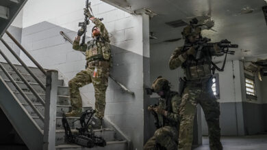 Soldiers conduct training inside a damaged building, moving up a stairwell with rifles raised while an AeroVironment ground robot supports their indoor clearing exercise.