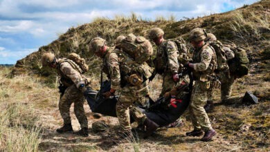 UK soldiers in full combat gear carry a casualty on a stretcher across grassy terrain during a blast-response training scenario