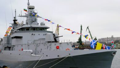 A modern grey Bulgaria warship docked at a harbor, decorated with colorful ceremonial naval flags and featuring a visible gun turret and radar mast against a cloudy sky