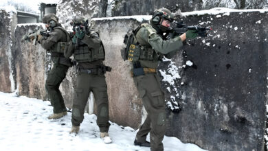 Soldiers in winter gear conduct live training behind a snow-covered concrete barrier, aiming their rifles while advancing in a coordinated formation