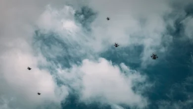 A drone swarm flies against a backdrop of partially cloudy, blue sky. The mood is technological and futuristic, with drones scattered at varying heights.