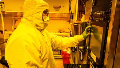 Technician wearing full cleanroom protective gear operates advanced quantum fabrication equipment inside a yellow-lit semiconductor cleanroom, interacting with a control panel on a precision processing system