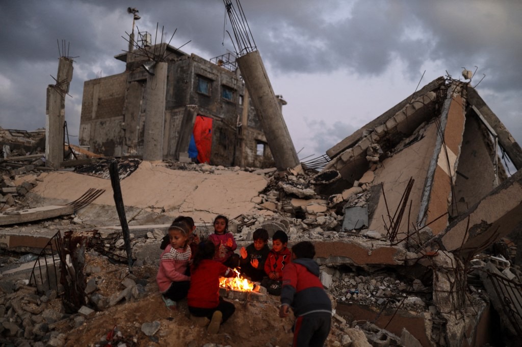 Displaced Palestinians warm up by the fire after their homes are destroyed following heavy rain in Gaza City, Palestine, on January 14, 2026.
