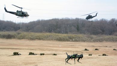 Robot dog unmanned ground vehicle moves across an open training field during a military exercise, with soldiers prone on the ground and two helicopters flying low overhead in the background