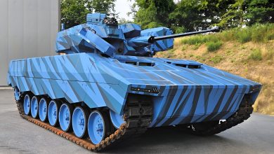 Condor tank on display, a modern tracked armored vehicle with angular blue camouflage, turret-mounted cannon, and multiple road wheels, parked outdoors near a facility