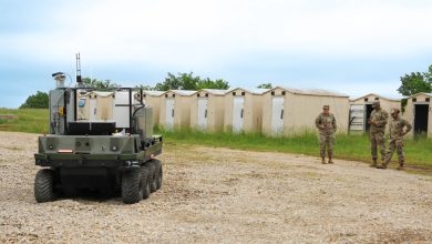 Military personnel observe autonomous ground robots operating in a field near temporary structures during a training or testing exercise.