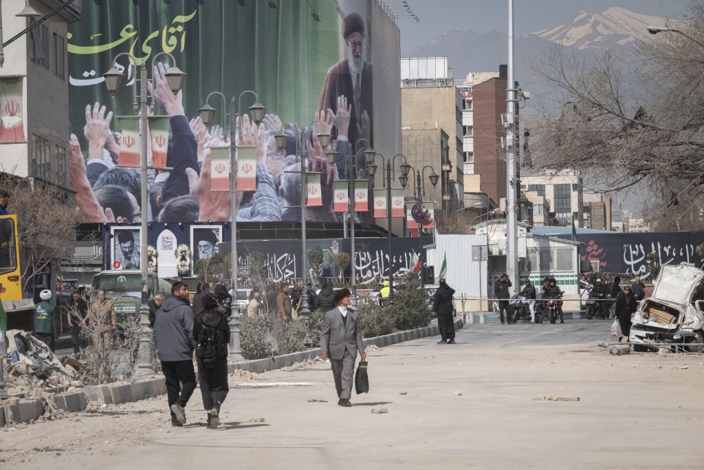 Iranian people walk through the area under a portrait of Iran's Late Leader, Ayatollah Ali Khamenei, outside a police facility that is completely destroyed during a US-Israeli military campaign in Tehran, Iran, on March 4, 2026.