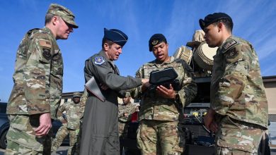 A US Air Force pilot officer and several airmen in OCP uniforms huddle around a handheld tactical device near field equipment, illustrating collaborative efforts to solve emerging drone warfare challenges